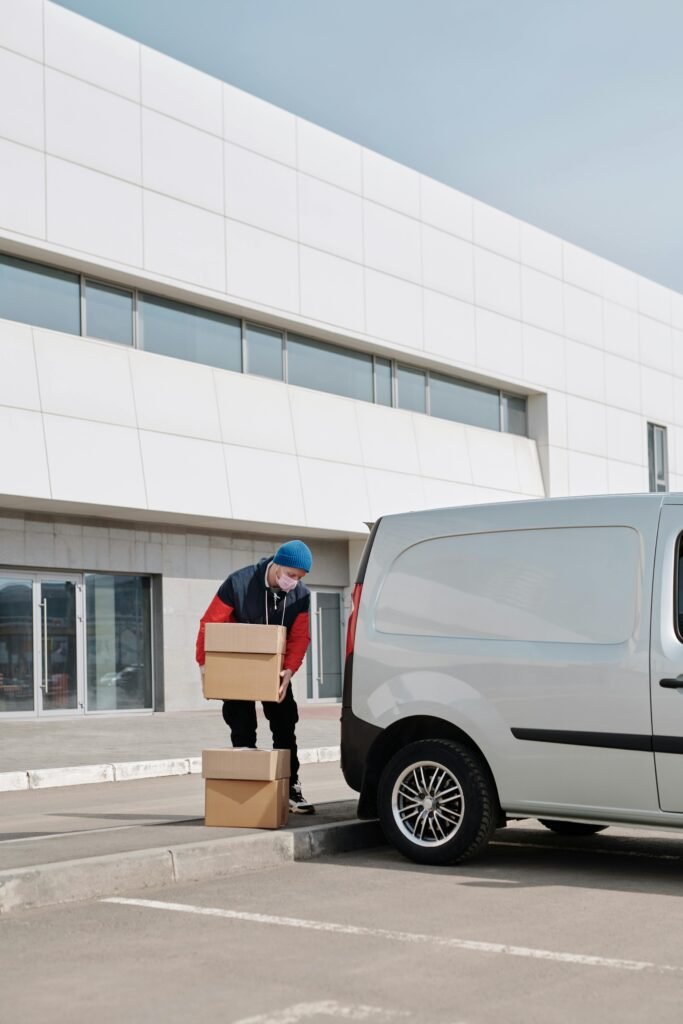 A delivery worker in a mask unloading boxes from a van in front of a modern building.