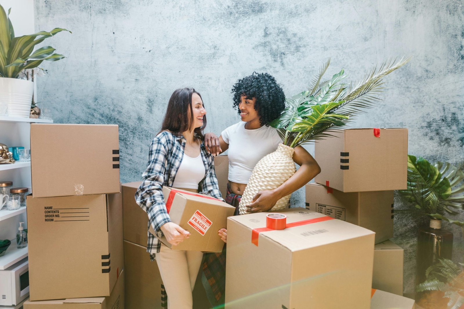 Two women smiling while unpacking in their new home with houseplants and moving boxes.